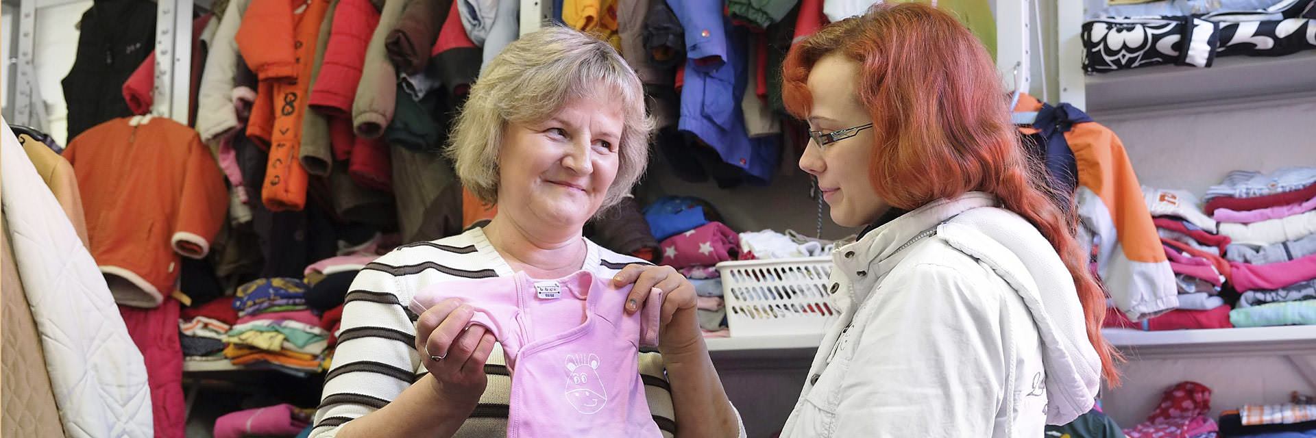 Ehrenamtliche Helferin in der Kleiderkammer der Tafel Stralsund überreicht gespendete Baby-Kleidung an eine Kundin. Im Hintergrund sind Regale mit sortierter Kleidung für Bedürftige.