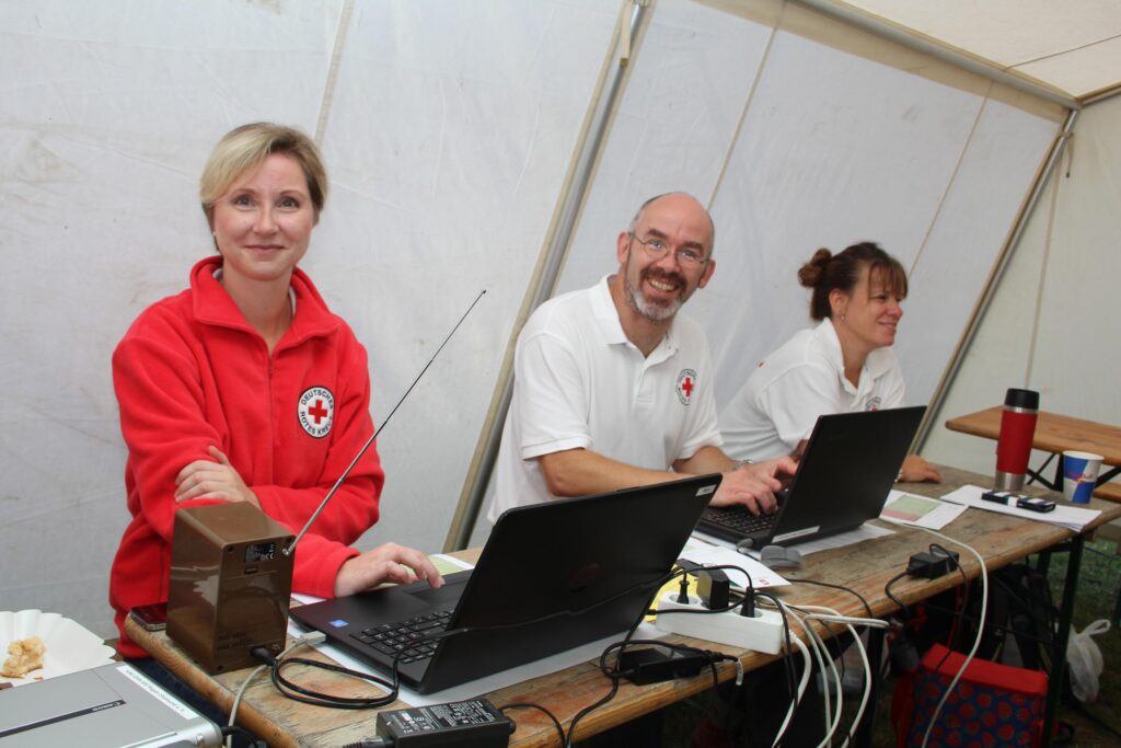 DRK-Suchdienst-Team in einem mobilen Einsatzbüro. Drei ehrenamtliche Helfer arbeiten an Laptops in einem Zelt, um vermisste Personen zu registrieren und Informationen bereitzustellen.