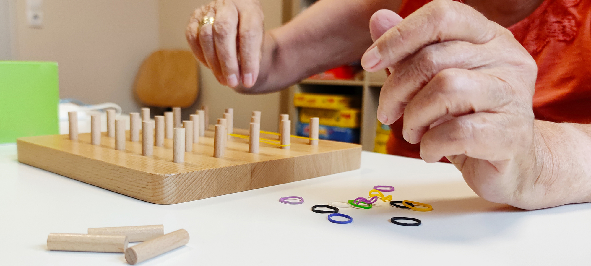 DRK-KV Rügen-Stralsund e. V. Ergotherapie: Maßnahme in der Ergotherapie. Beweglichkeits- und Geschicklichkeitstraining mit der Hand. Hierbei werden Gummis über Daumen und Zeigefinder gespannt auf Holzstifte gezogen.