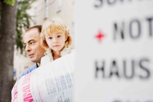 Ein Mann hält ein kleines blondes Mädchen, das ruhig in die Kamera blickt. Im Vordergrund ist unscharf ein Schild mit dem roten Kreuz