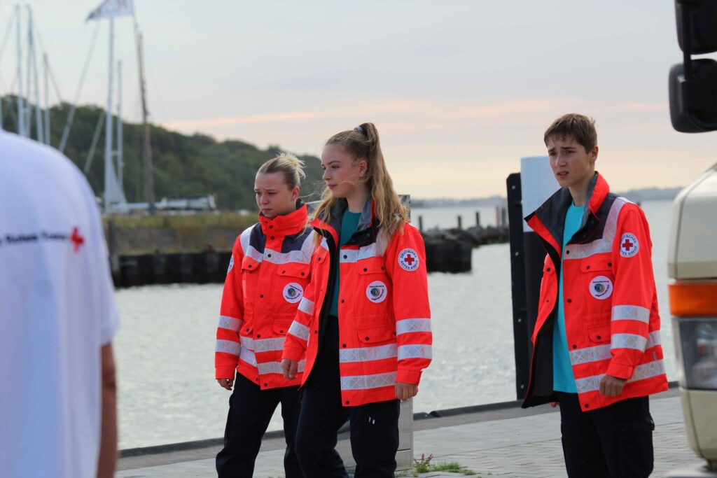Drei junge Mitglieder des Jugendrotkreuzes (JRK) in leuchtend orangen Einsatzjacken mit DRK-Logo stehen an einer Hafenpromenade. Sie wirken aufmerksam und konzentriert.