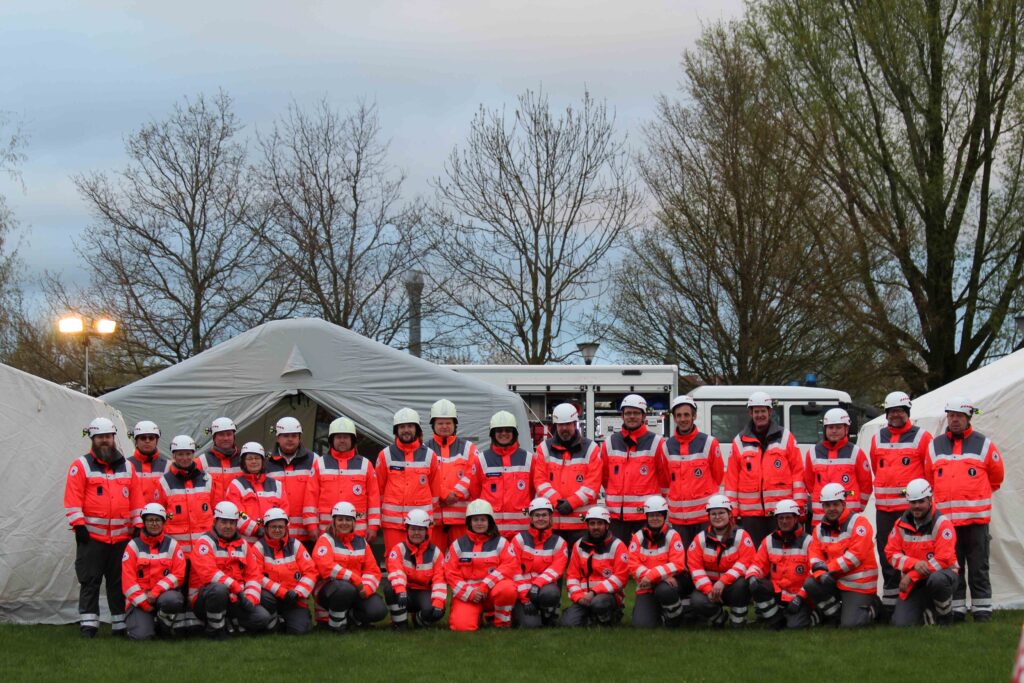 Dieses Bild zeigt eine Gruppe von DRK-Einsatzkräften in orangener Schutzkleidung mit Helmen. Sie stehen vor Notfallzelten und Einsatzfahrzeugen in einem Freigelände.