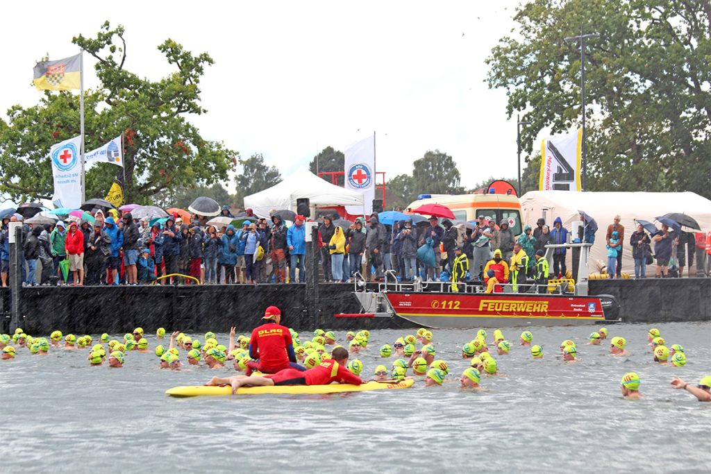 Schwimmerinnen und Schwimmer starten beim 26. Internationalen Vilm-Schwimmen im Lauterbacher Hafenbecken bei Regen und Wind, abgesichert von Einsatzkräften an Land und auf dem Wasser.